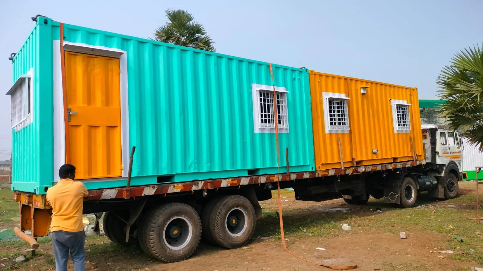 Two porta cabins in turquoise and orange, strapped side by side on a flatbed truck, with barred windows and a panelled door, and a worker standing at the trailer edge