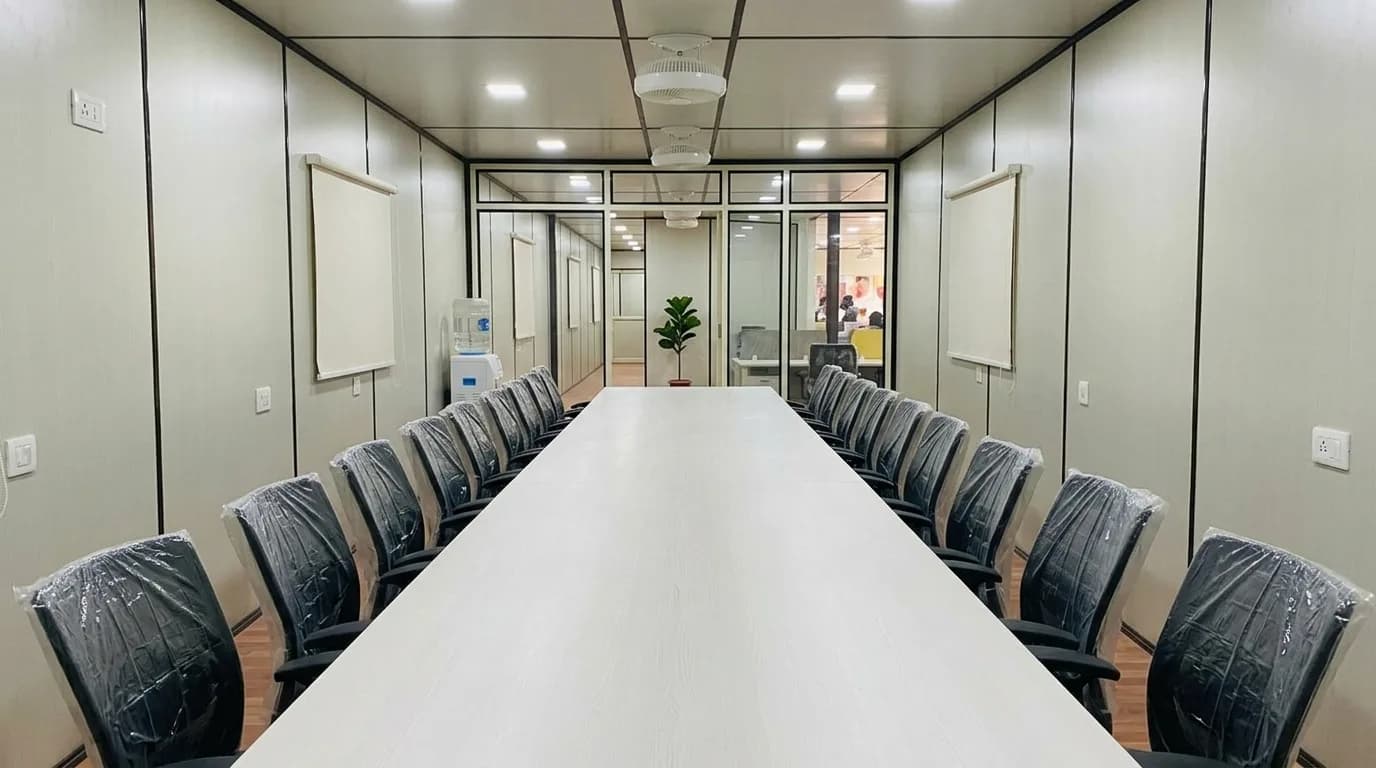 Meeting room inside a prefabricated office with a long conference table, chairs still in factory wrapping, ceiling fans, recessed lighting and a glazed partition to an open-plan area