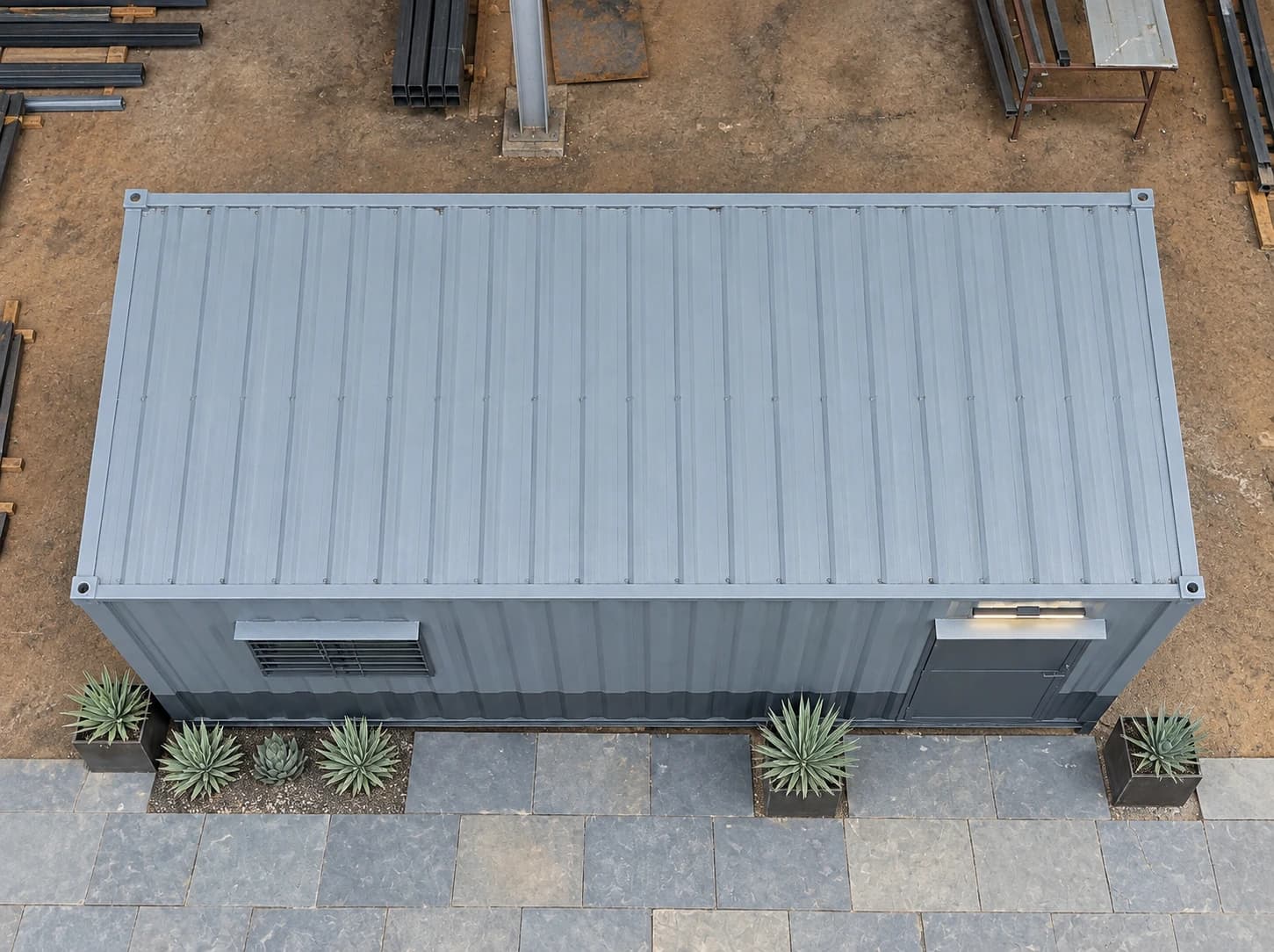 MS porta cabin seen from above, showing its corrugated roof, a barred window and a door with a light on its long side, and planters along a paved strip, with steel sections stacked on the ground nearby
