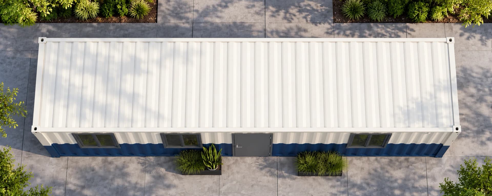 Long office cabin seen from directly above, with a white corrugated steel roof, a navy lower band along the front, a grey door, three windows and two planters on a paved yard