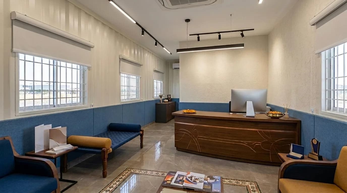 Reception area inside a prefabricated cabin with a curved timber desk, a monitor, armchairs and a bench for waiting visitors, air conditioning and barred windows along one wall